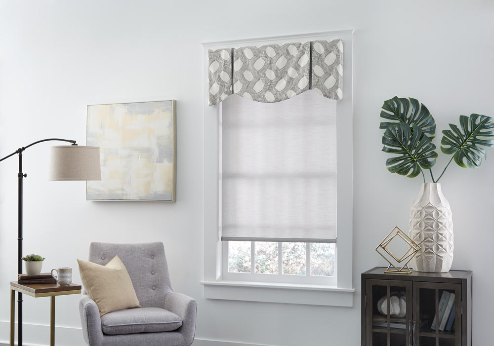 gray Genesis® Roller Shade with an Interior Masterpieces® gray and white patterned fabric valance next to a light gray chair with a tan pillow on it