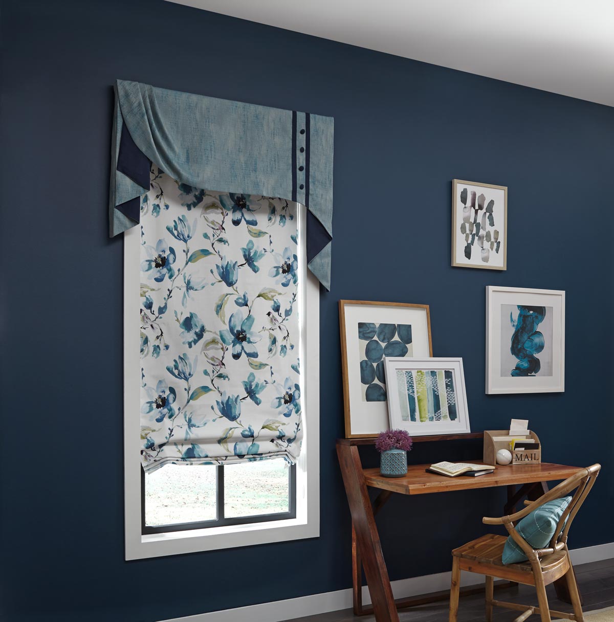 Window in a blue wall with an Interior Masterpieces® custom fabric cornice and a roman shade next to a wooden table with paintings hung behind it