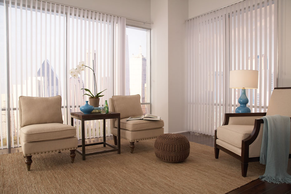 Two large picture windows with Discoveries® Vertical Blinds hanging in them in the open position in a room with tan furniture and blue accent pieces