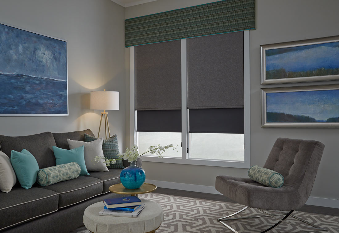 Two large gray Genesis® Roller Shades with a green and yellow striped Interior Masterpieces® Fabric Cornice spanning both shades and a dark gray couch with accenting blue, gray and green Custom Pillows