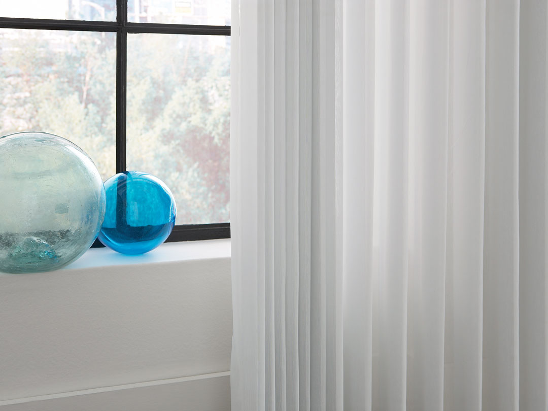 Close up of white Sheer Visions® Vertical Blinds with some blue glass decorations in a window