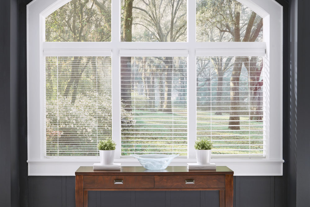 three white Heartland Woods® Wood Blinds in 1", 2" and 2 1/2" slat sizes in the open position behind a brown desk against dark gray walls