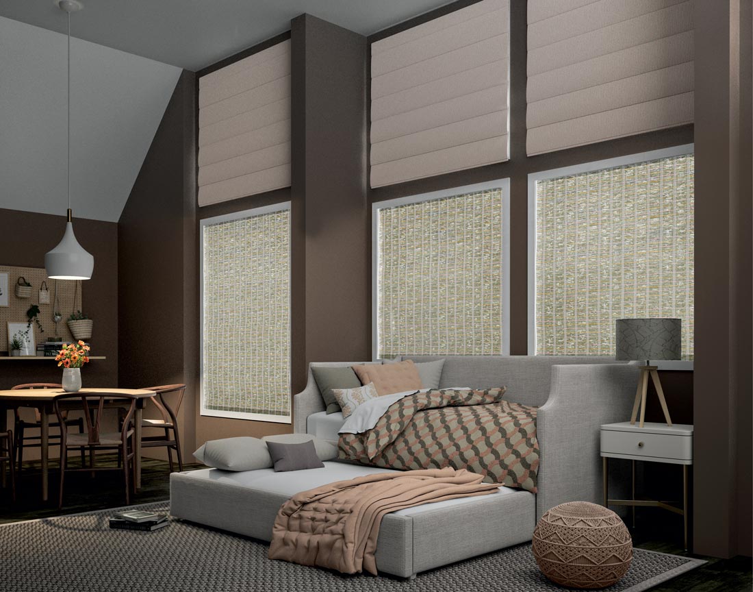 Light tan Interior Masterpieces® Motorized Roman Shades in three windows high near the ceiling above 3 windows with Manh Truc® Motorized Woven Wood Shades behind a couch and sleeper with Interior Masterpieces® Custom Blankets, Pillows and Throws