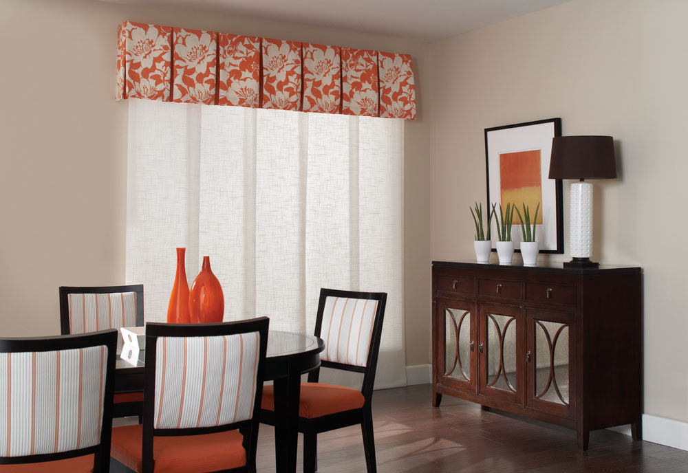White Genesis® Panel Track system in a large window in a dining room with an Interior Masterpieces® ornage and white fabric wrapped cornice with a black table and orange decorations on it