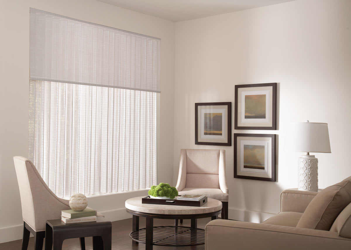 A large, closed white Manh Truc® Woven Wood Panel Drape in a picture window with a Valance in the same material next to a tan table and chairs