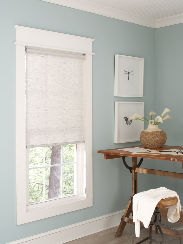 white Genesis® Custom Roller Shade in a room with light blue walls and a brown desk nearby