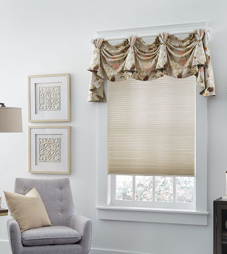 A light taupe cellular shade hangs in the window of a seating area featuring a modern light gray chair, and a floral fabric swag pole valance.