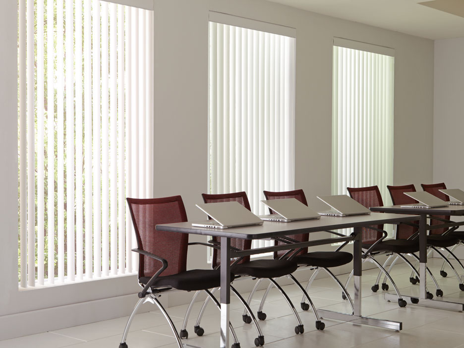 3 large windows with white colored Discoveries® Vertical Blinds in them in an office with several red chairs and laptops