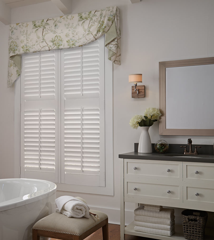 white Parke® Shutter with a tan and yellow floral patterened Interior Masterpieces® Fabric Valance in a bathroom with beige walls and a white stand alone tub in the foreground