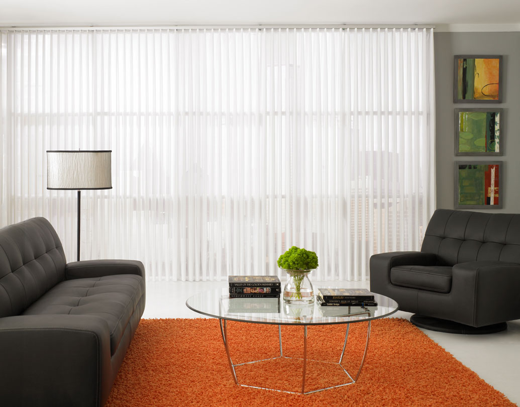 White Sheer Visions® Vertical Blinds hanging in a large window in a room with an orange rug and black furniture