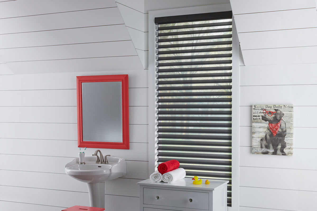 A black Tenera® Sheer Shade with vanes open in a white bathroom with a sink and red mirror next to it