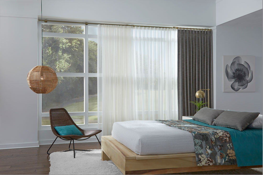 Bedroom scene with a large picture window and a sheer white Interior Masterpieces® Drapery with a Gray Solid stationary panel next to a bed with Custom Interior Masterpieces® Pillows and Blankets in Teal and a Floral pattern