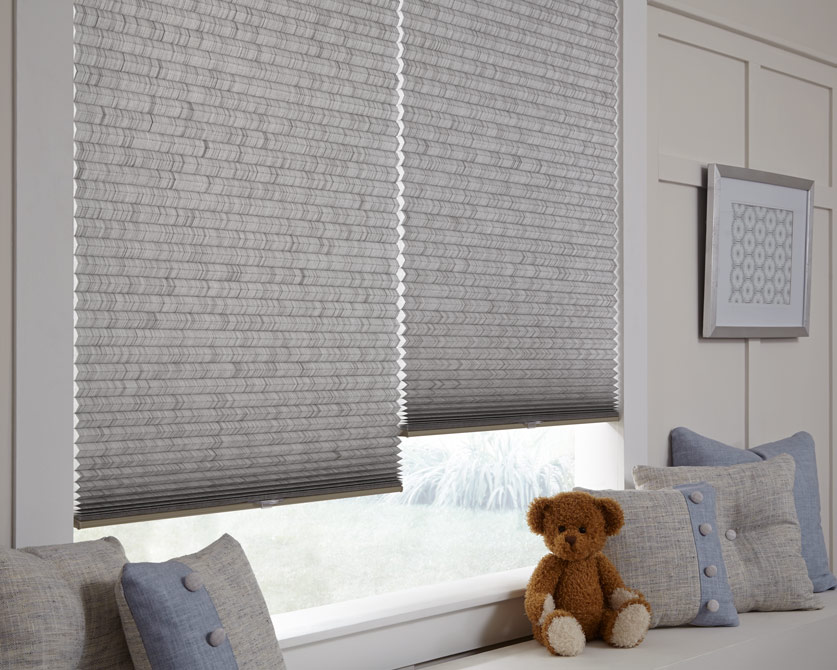 Two child-safe textured gray cellular shades hang in a cozy window nook, with custom throw pillows and a teddy bear on the window bench beneath.