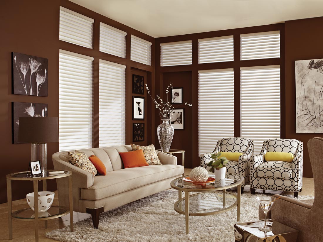 white Tenera® motorized sheer shades set against a brown wall with a tan couch and chairs that have Interior Masterpieces® custom pillows on them