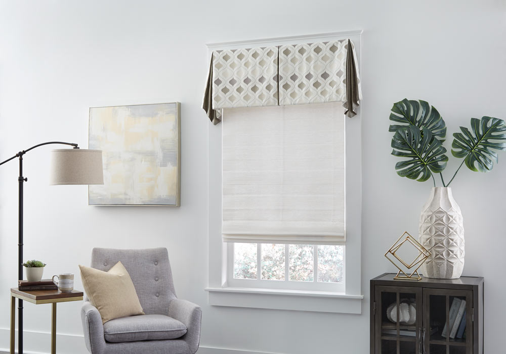 gray Genesis® Roller Shade with Interior Masterpieces® Fabric Valance with a tan diamond pattern next to a light gray chair with a tan pillow on it