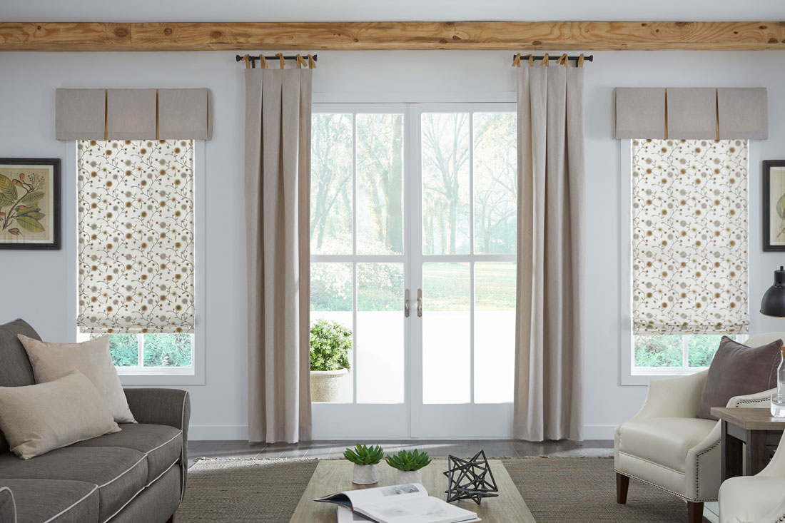 two cream colored floral patterned Interior Masterpieces® Fabric Shades with custom tan Fabric Wrapped valances hanging on each side of a patio door with tan Draperies