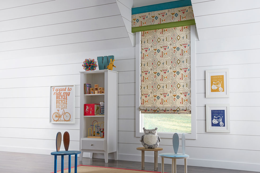 tan Interior Masterpieces® fabric shade with geometric pattern and accenting fabric wrapped cornice in a child's room with small chairs and playthings on shelves