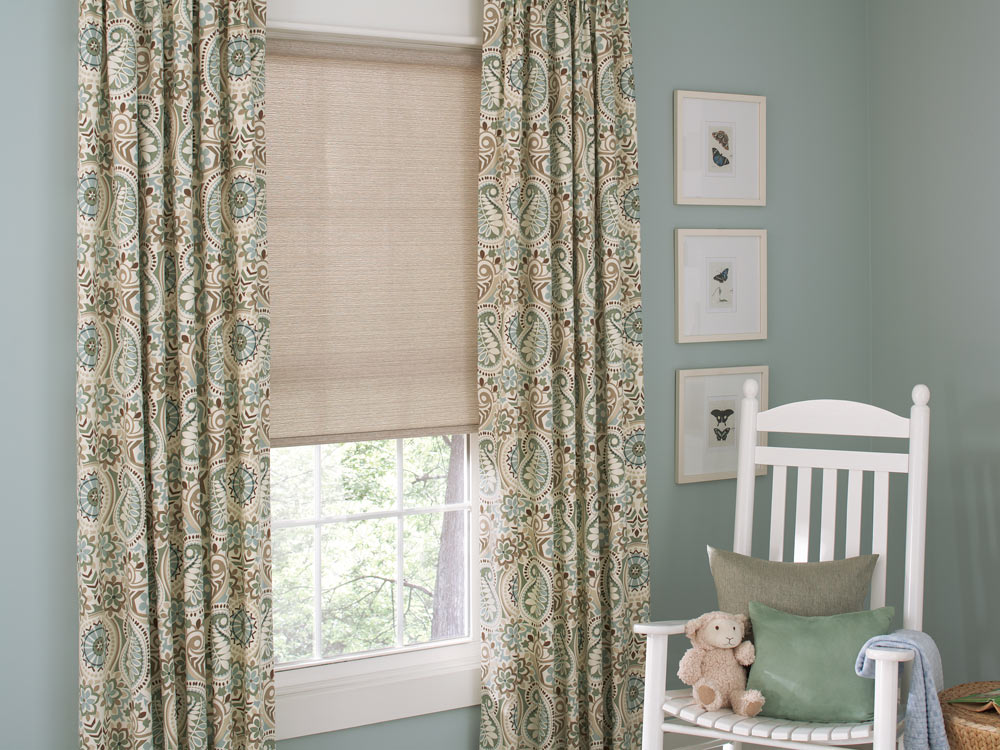 tan Genesis® Roller Shades and floral patterned Interior Masterpieces® Draperies against blue walls with a white chair nearby and a stuffed animal on it
