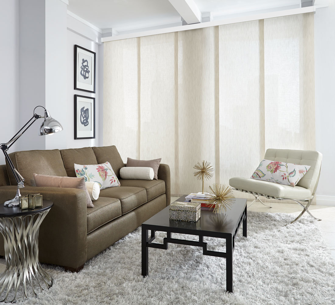 Very wide window with a city skyline in the background with a light tan Genesis® Panel Track hanging in front of it with a dark couch and white chair that has custom pillows on them