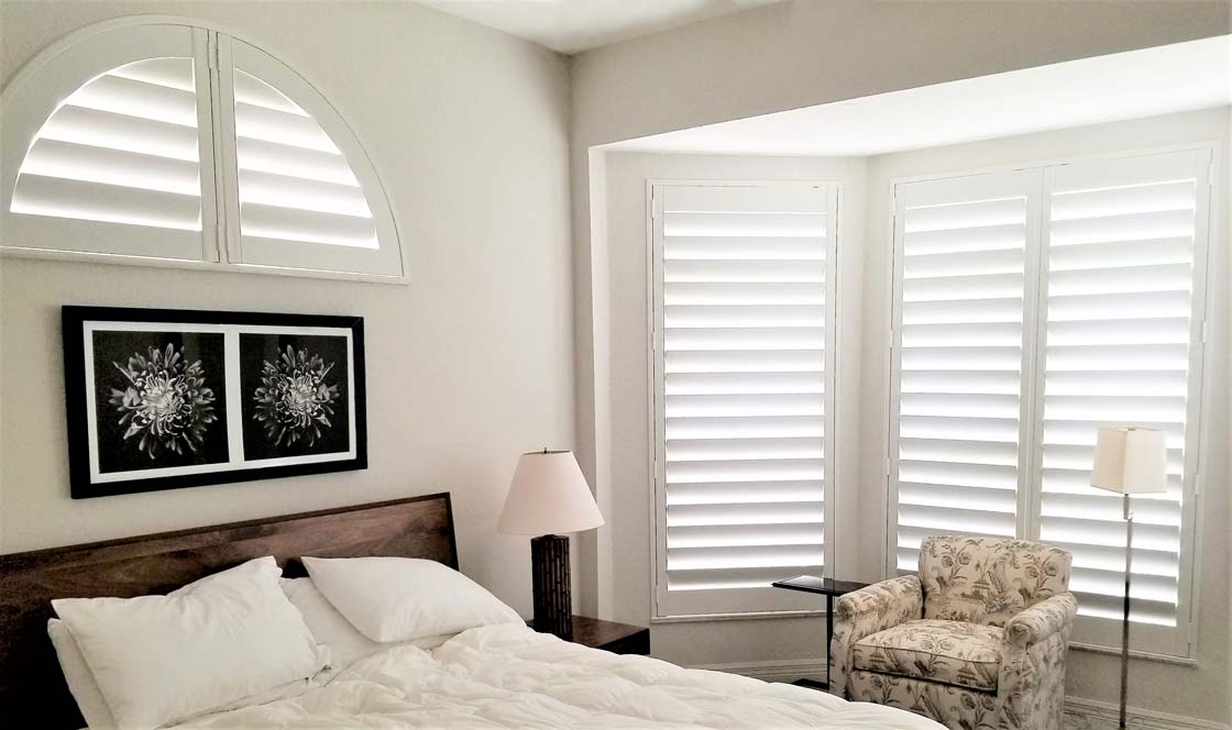 Bedroom with Parke® Shutters in a bay window and an arched window above a bed