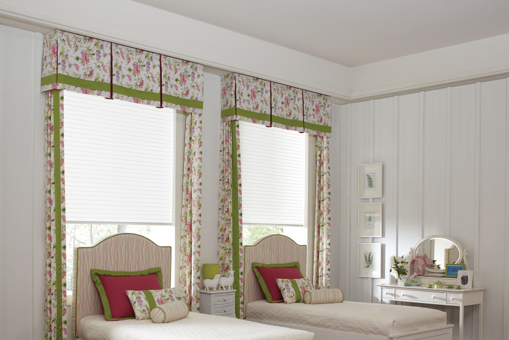 Two white Tenera® Sheer Shades with Interior Masterpieces® Fabric Cornices and Draperies in a floral pink, green, purple, and white pattern with matching Headboard and Custom Bedding