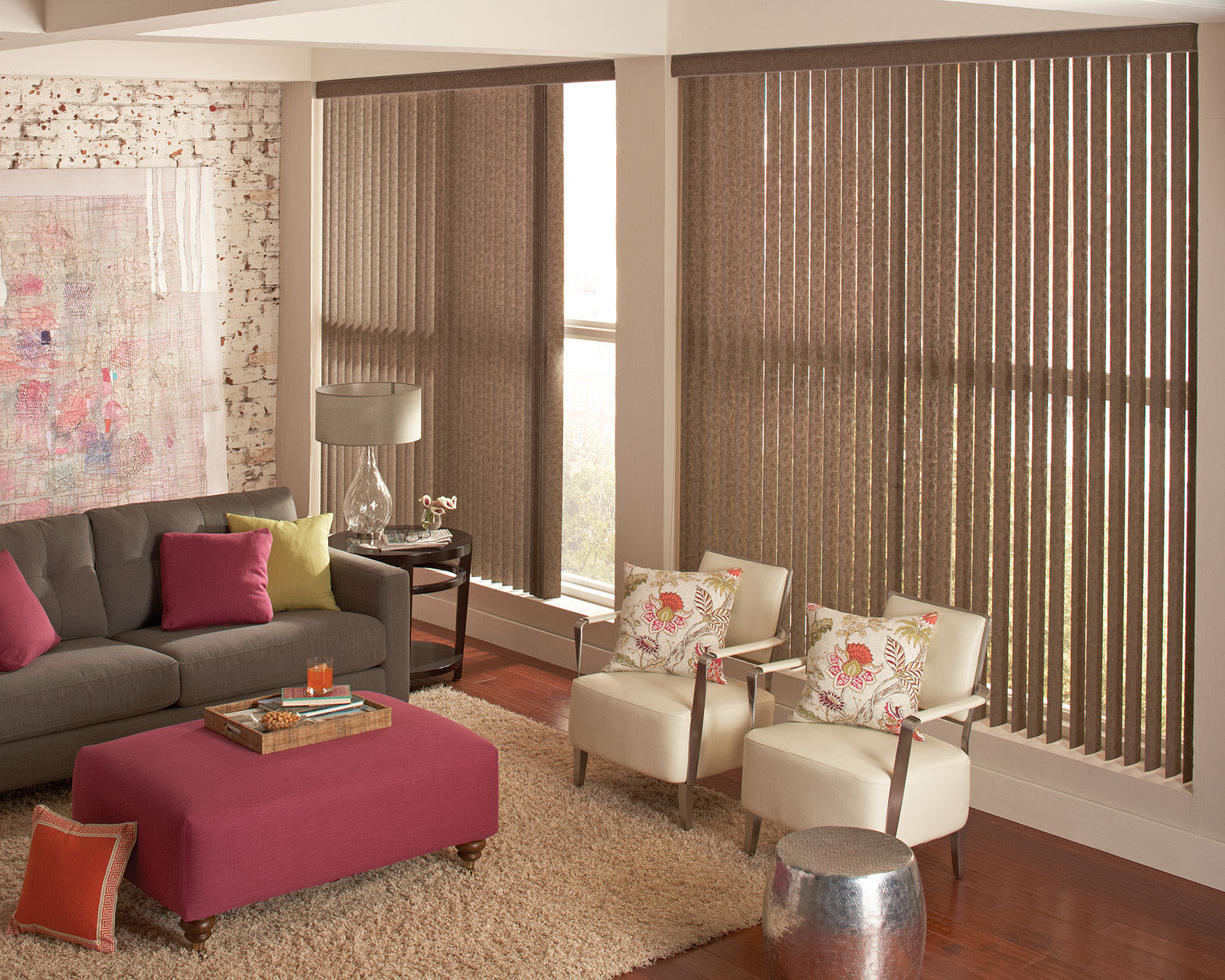 Textured taupe vertical blinds hang in an exposed brick loft apartment with custom throw pillows in bright colors of fuchsia and green.