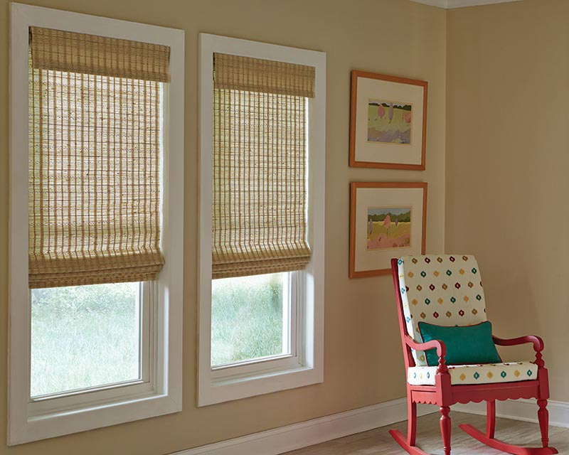 Three cornsilk colored child-safe flat roman shades hang in a tan room with a red rocking chair.