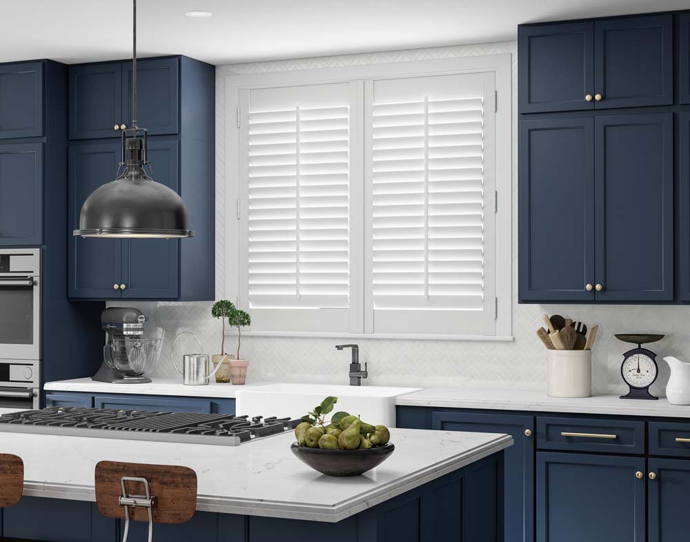 white Parke® Shutters in a kitchen with blue cabinets, a sink, and an island stove top with marble counters in the foreground