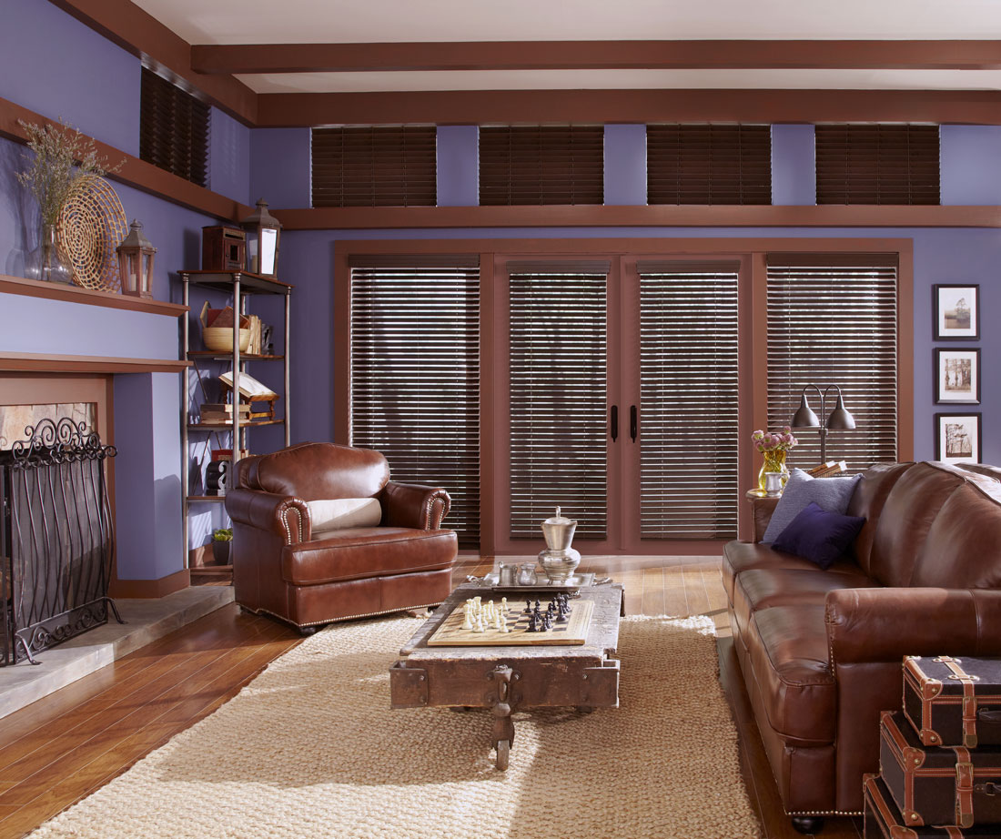 several dark brown Heartland Woods® Wood Blinds with Valances against a purple wall with brown lather furniture in the room