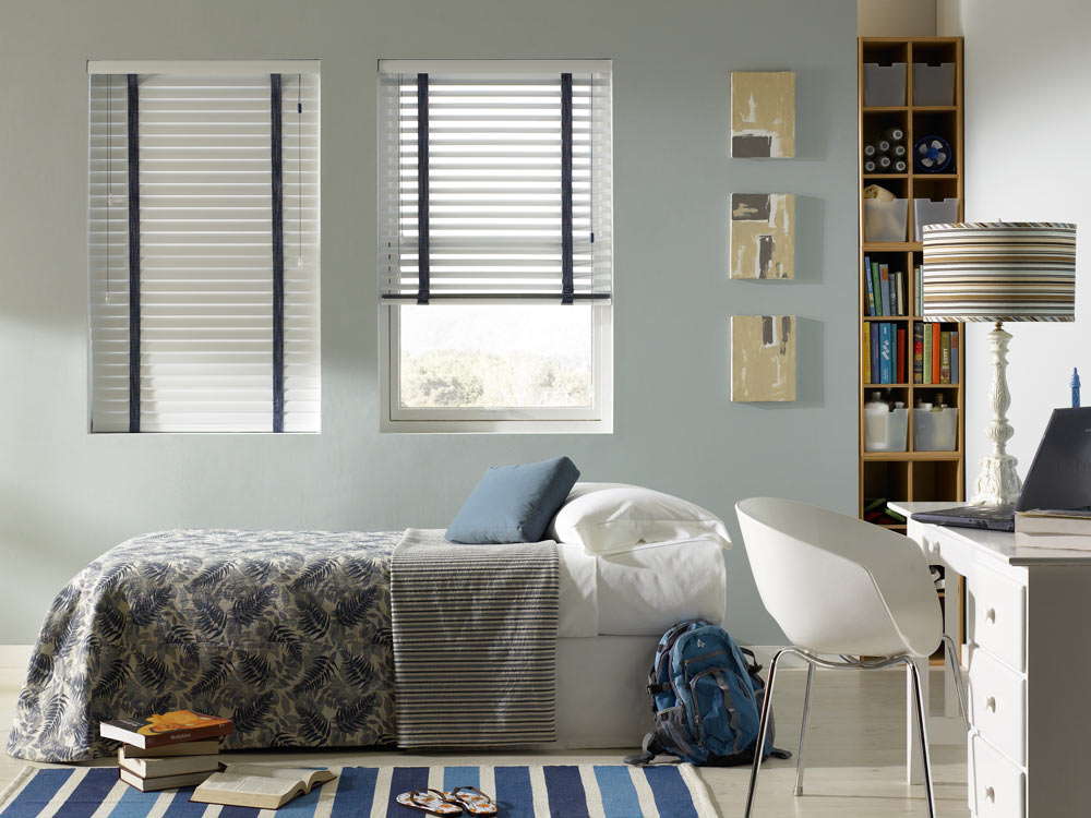 Two white Classic Collection® Aluminum Blinds with wide blue Decorative Tape behind a bed with white sheets and blue and gray custom bedding