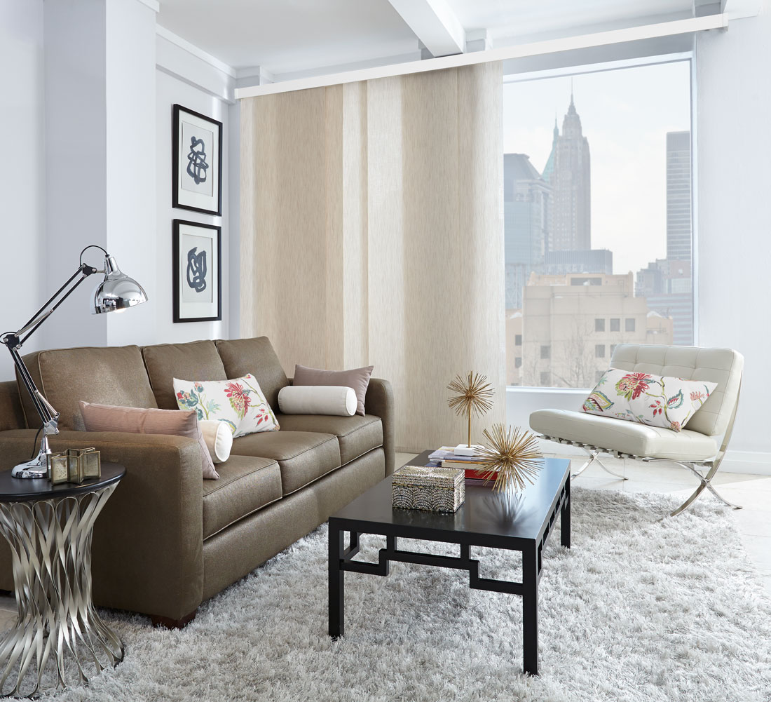 Very wide window with a city skyline in the background with a light tan Genesis® Panel Track hanging in front of it with a dark couch and white chair that has custom pillows on them