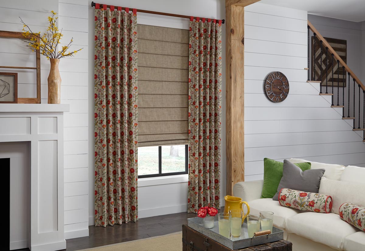 Tan roman shade in a window with tan draperies with red flowers hanging on each side in a white room with a couch and custom draperies pillows