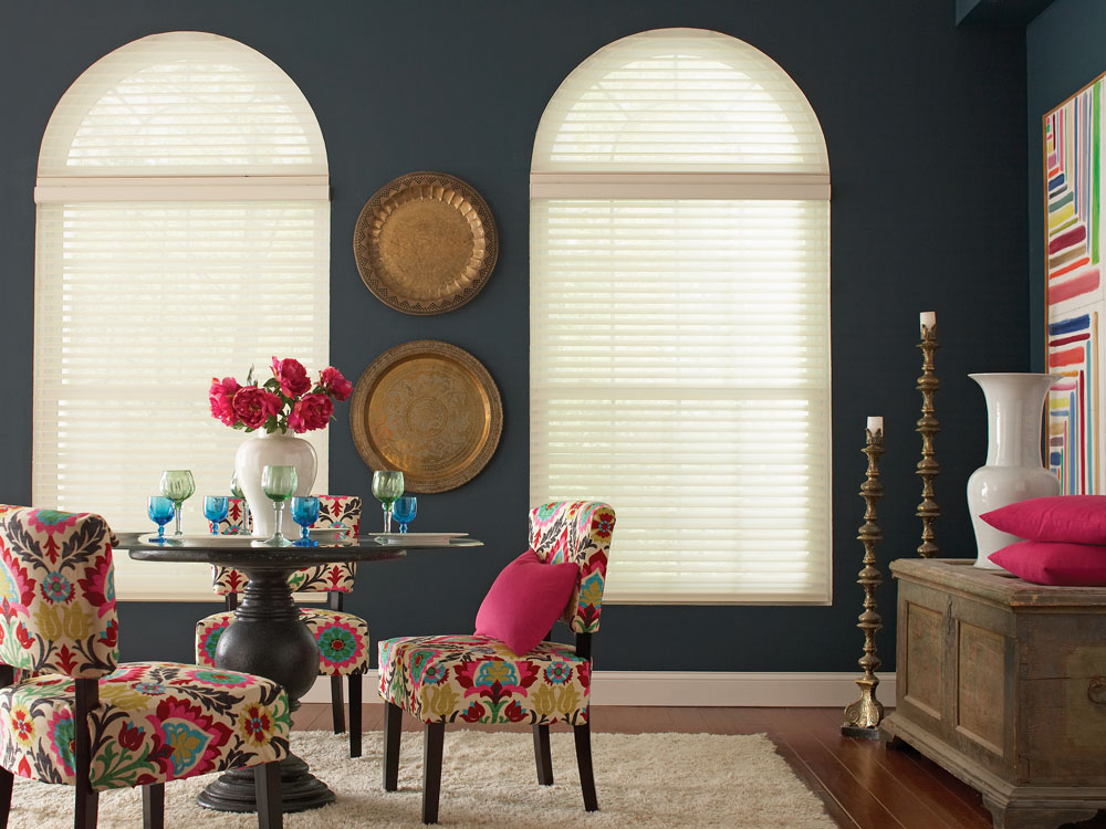 Two lightly colored Tenera® sheer shades with an arch top set against a dark blue wall with a table that has brightly colored flowers and cups on it and chairs that have Interior Masterpiece® pillows on them