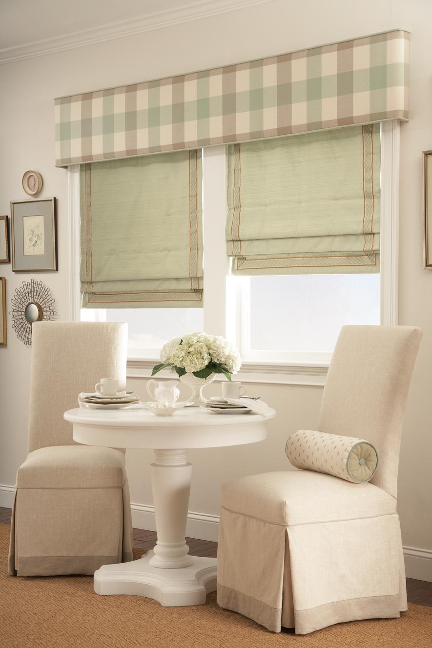 Two windows with Green Interior Masterpieces® Hobbled Roman Fabric Shades with light brown trim and a tan Custom Cornice spanning the width of both windows with a brown and green striped pattern on it behind two small chairs and a table with white flowers