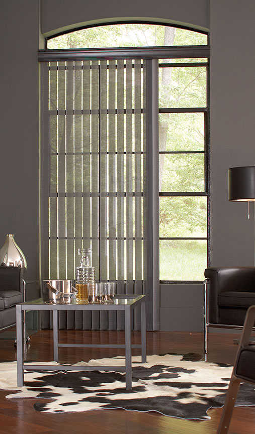 Semi-Sheer charcoal vertical blinds hang on a carved wood valance in a modern room featuring leather chairs with metal accents and a cowhide rug.