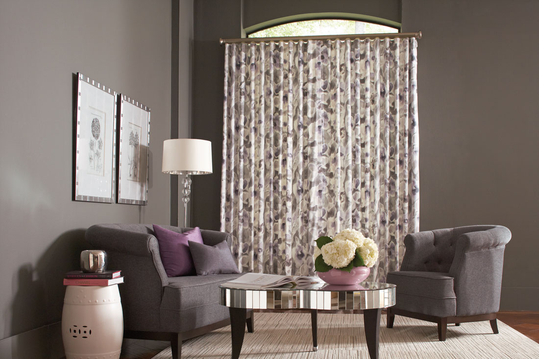 A white Interior Masterpieces® Drapery with a purple floral pattern in a window with a gray couch and chair that hasInterior Masterpieces® Custom Pillows in purple and gray