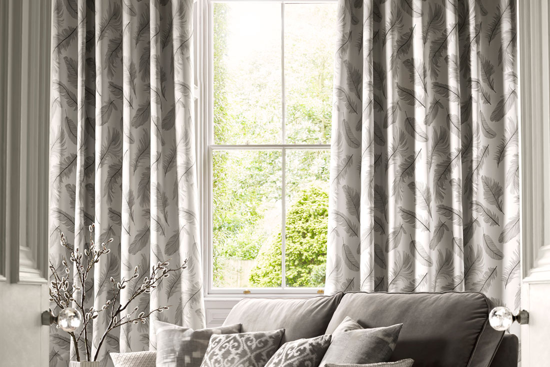 light gray colored pattern of feathers on Interior Masterpieces® Drapes with a hanging in a window with a gray couch that has Interior Masterpieces® Custom Pillows