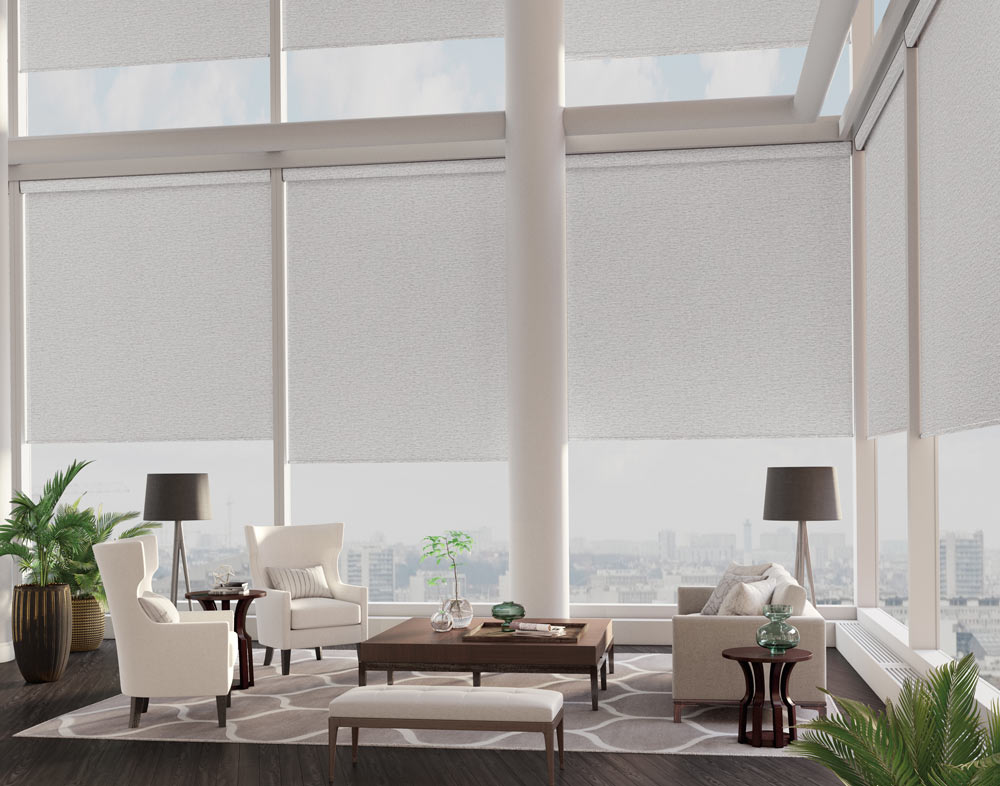 Several large tan Genesis® Motorized Shades in a room with big double stacked windows on either side and white furniture in the foreground