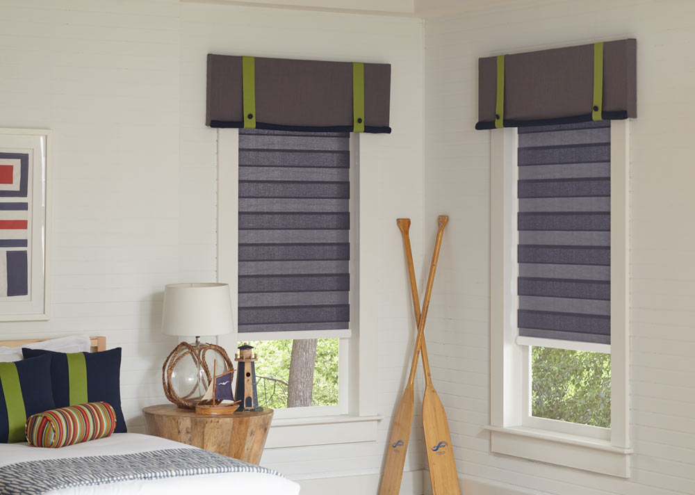 Two light gray Allure® Transitional Shades with Interior Masterpieces® Fabric Cornices with accenting green decorative tapes on them in a bedroom with oars leaning on the wall in between them