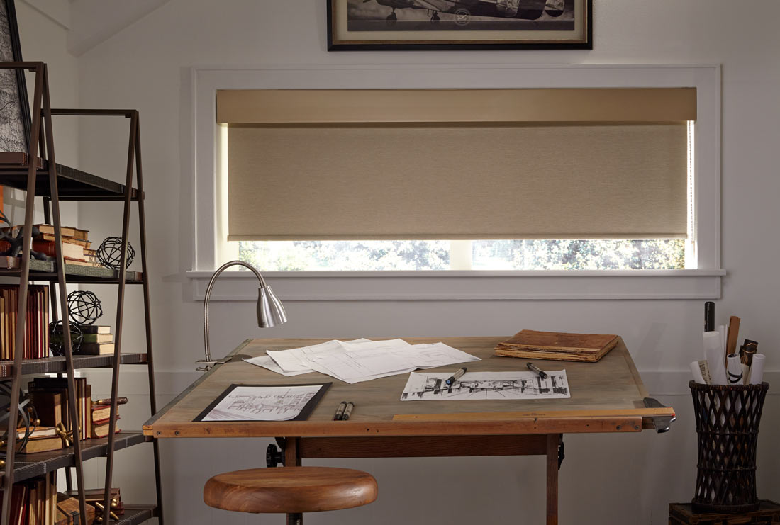 Wide and short brown Genesis® Roller Shades & Fabric Valance with matching material behind a wooden desk in a room with beige walls