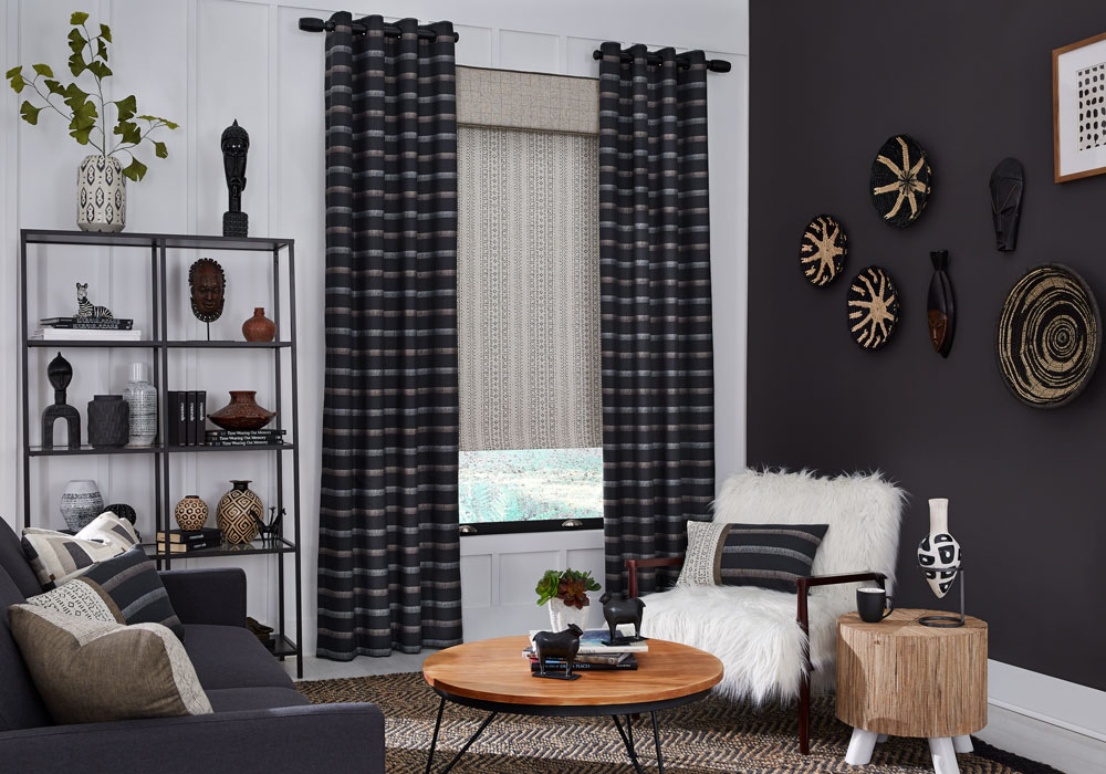 Tan and black patterned Interior Masterpieces® fabric shade with custom fabric cornice and dark draperies with white and gold stripes in a room with a fluffy chair, couch and custom pillows matching the draperies material