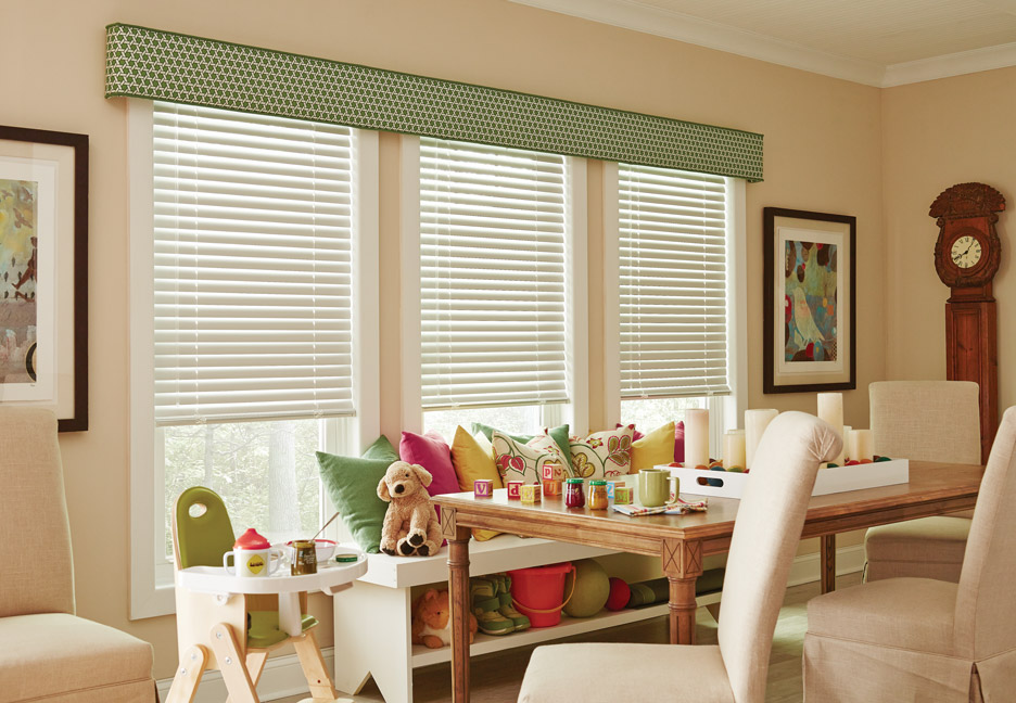 Three white Classic Collection® Aluminum Blinds with a green Interior Masterpeices® Fabric Cornice spanning across them and colorful Custom Pillows and kid's toys on a bench in front