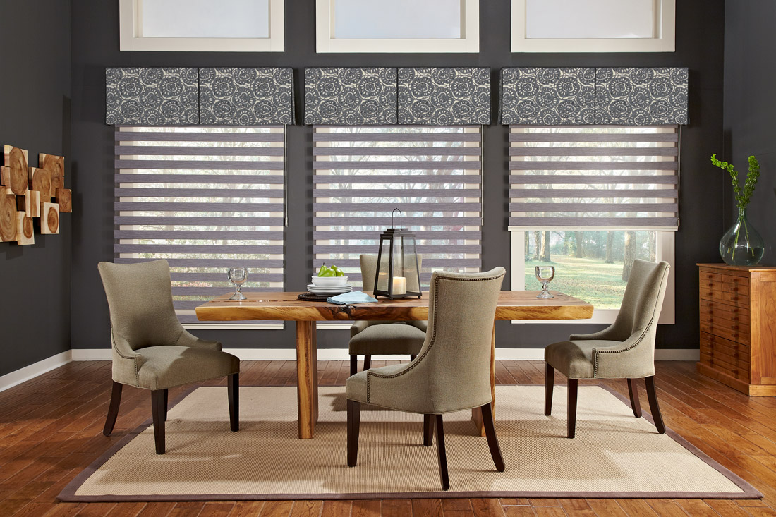 three large gray Allure® Transitional Shades with green and blue floral patterned Interior Masterpieces® fabric cornices against a dark gray wall behind a brown table and chairs