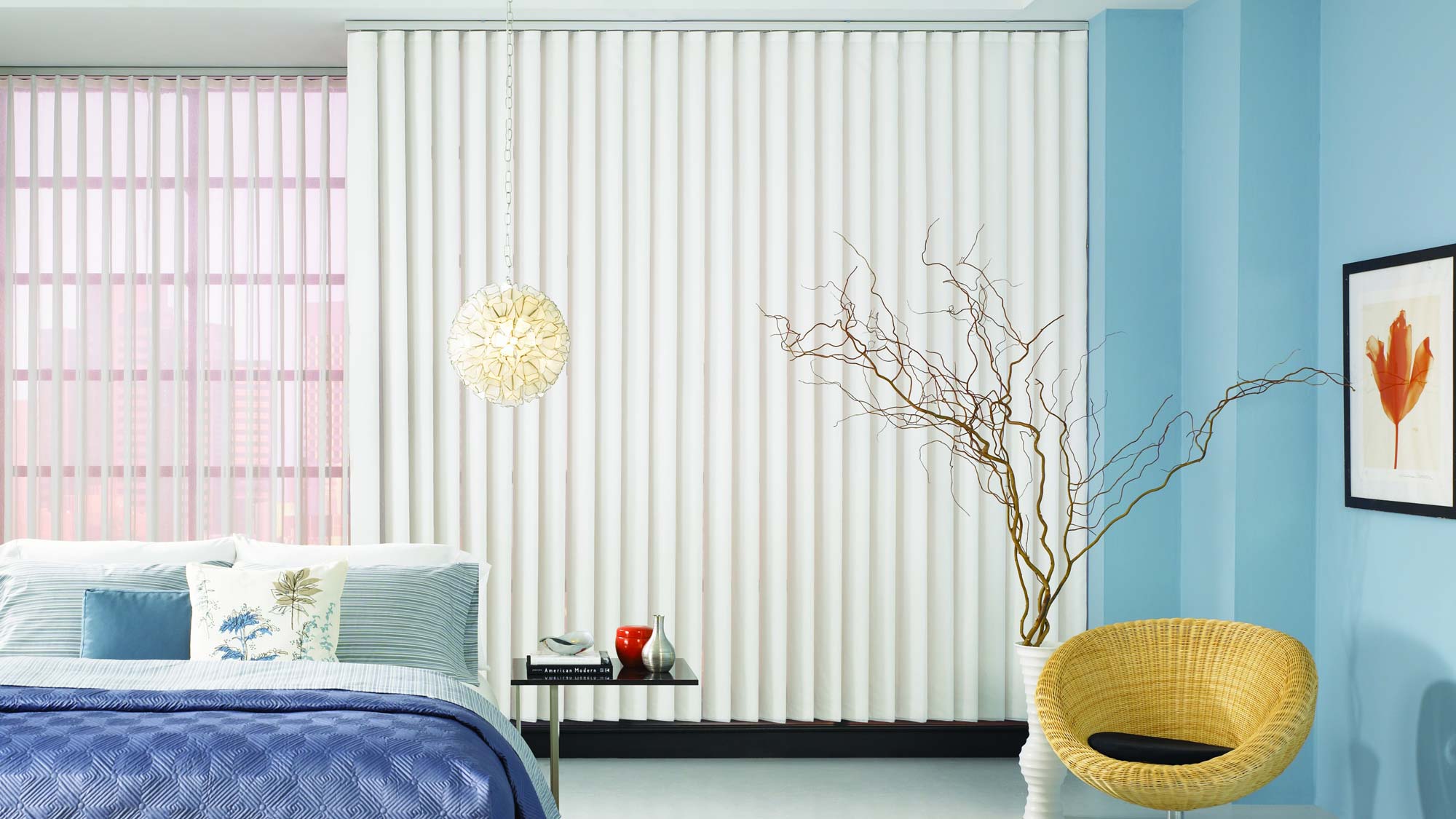 Vertical blinds and sheer fabric wrapped vertical blinds cover a large window in a room with blue walls and bedding.