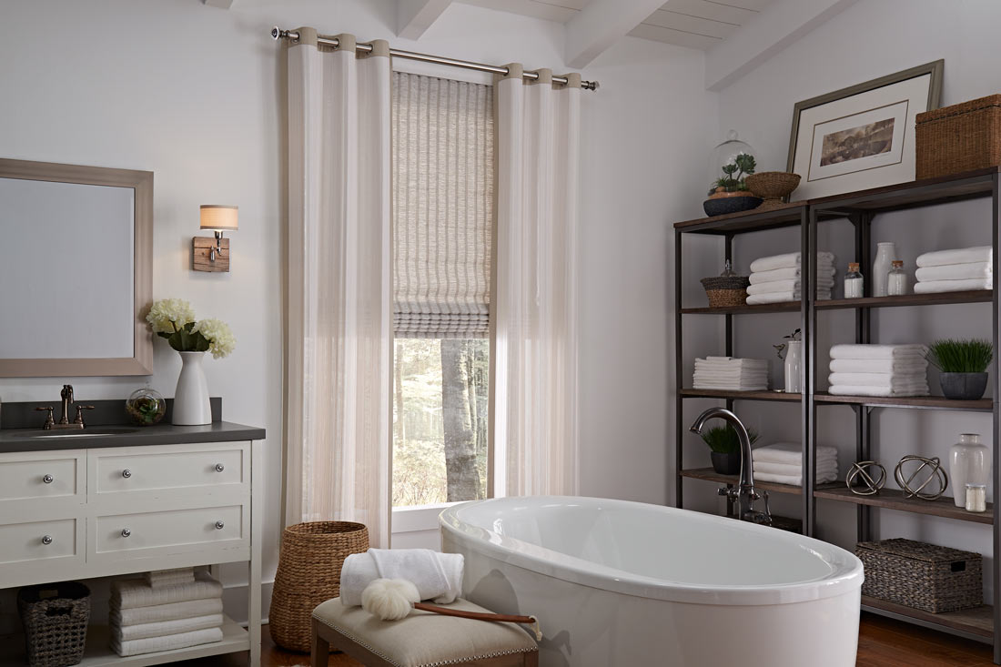 a tan Genesis® Hobbled Roman Shade with tan Manh Truc® Grommet Panel hanging on silver Custom Hardware in a bathroom behind a white stand alone tub next to shelves with towels