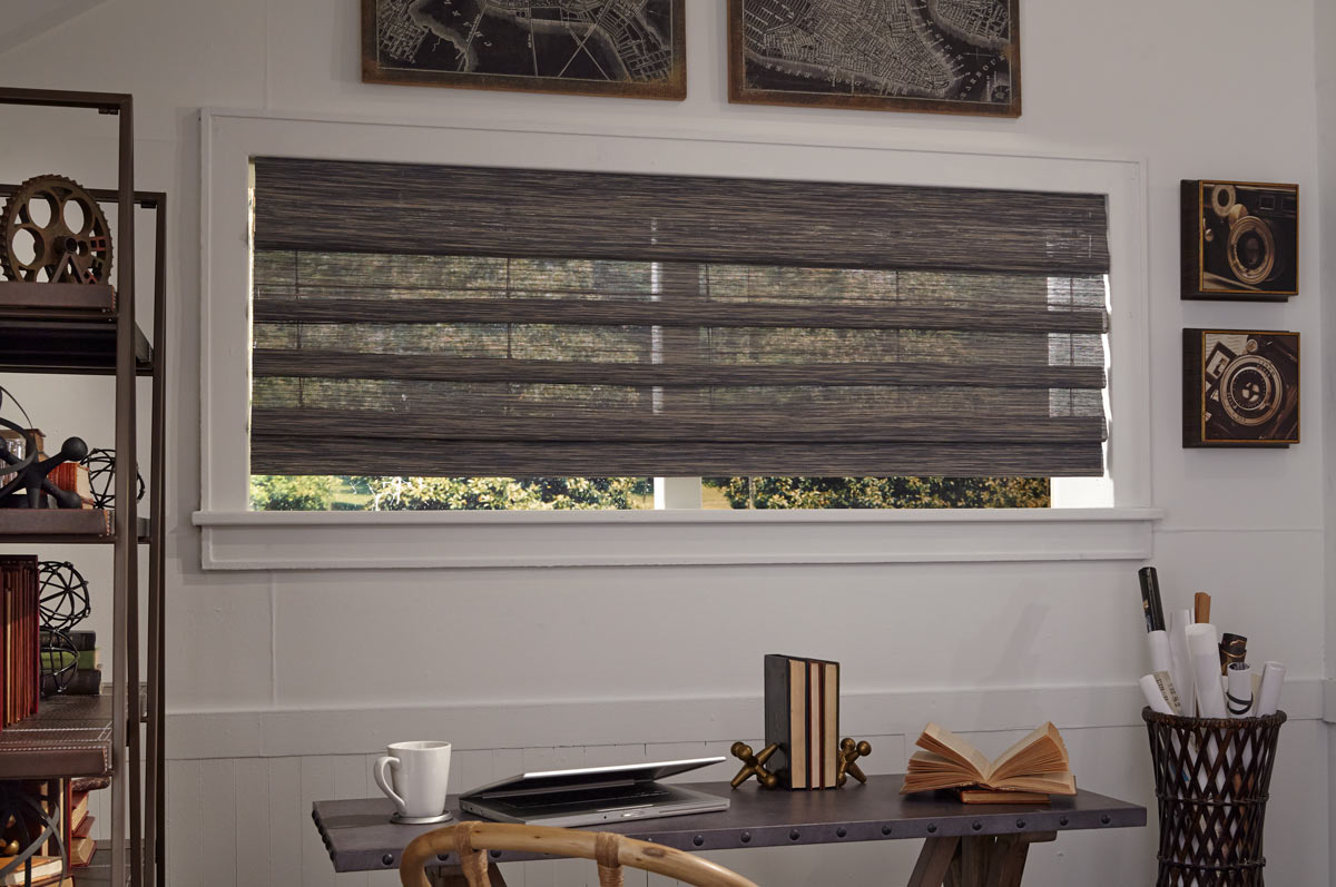 Wide and dark Manh Truc® woven wood shade behind a desk with pictures of maps in frames and a dark desk in the foreground