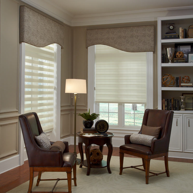 two cream colored Allure® Transitional Shades with brown Interior Masterpieces® Fabric Cornices in a room behind a small dark desk with matching chairs