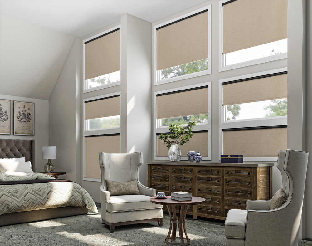 A grid of nine windows each with a tan Genesis® Motorized Roller Shade and a black headrail behind a large dresser and chairs