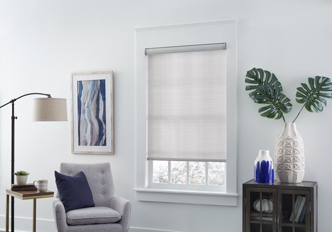 light gray Genesis® Custom Roller Shade next to a gray chair with a blue pillow and a blue painting on the wall