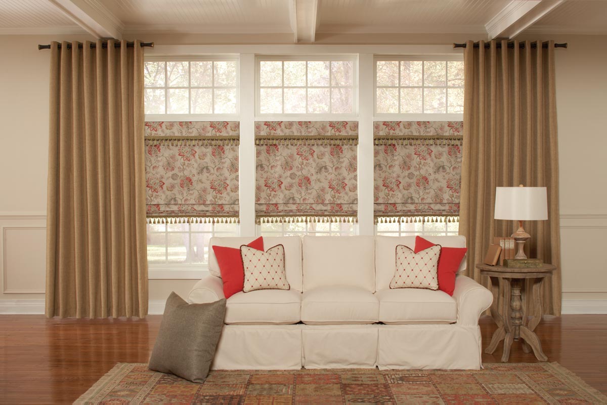 Brown colored Interior Masterpieces® draperies hanging with Custom Hardware on the outside of 3 windows that have Interior Masterpieces® Flat Roman shades that have a tan and floral pattern with Embellishment Trim on them behind a couch with Interior Mast